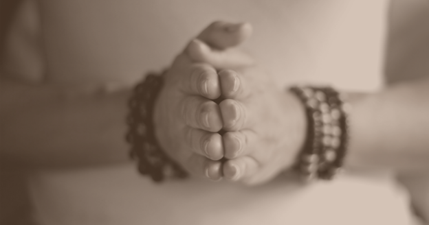 Close-up of hands in a prayer position, adorned with beaded bracelets, against a softly lit background.